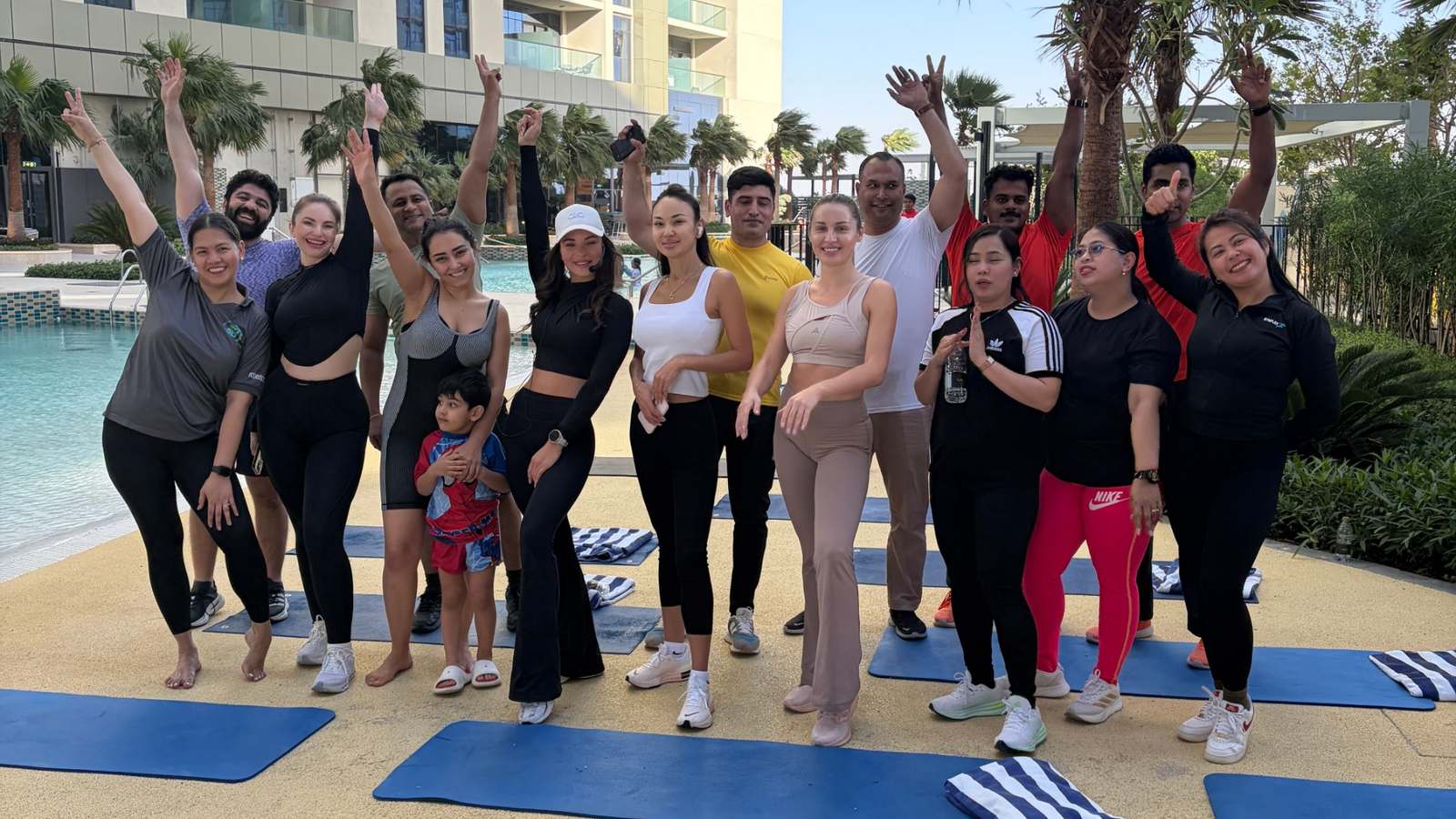A smiling group of people in sportswear waving their arms in the air after an outdoor Ellefit wellness session on exercise mats beside a pool in Dubai